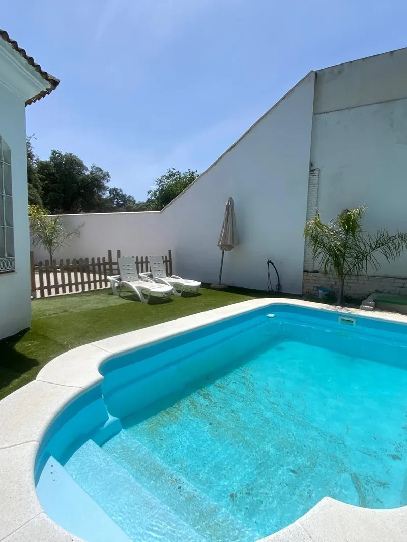 Borde de la piscina con la escalera de acceso, una sombrilla cerrada y el muro blanco al fondo