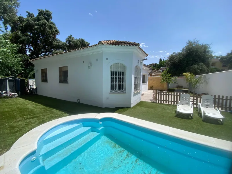Piscina en primer plano, con dos tumbonas sobre el césped y la casa al fondo